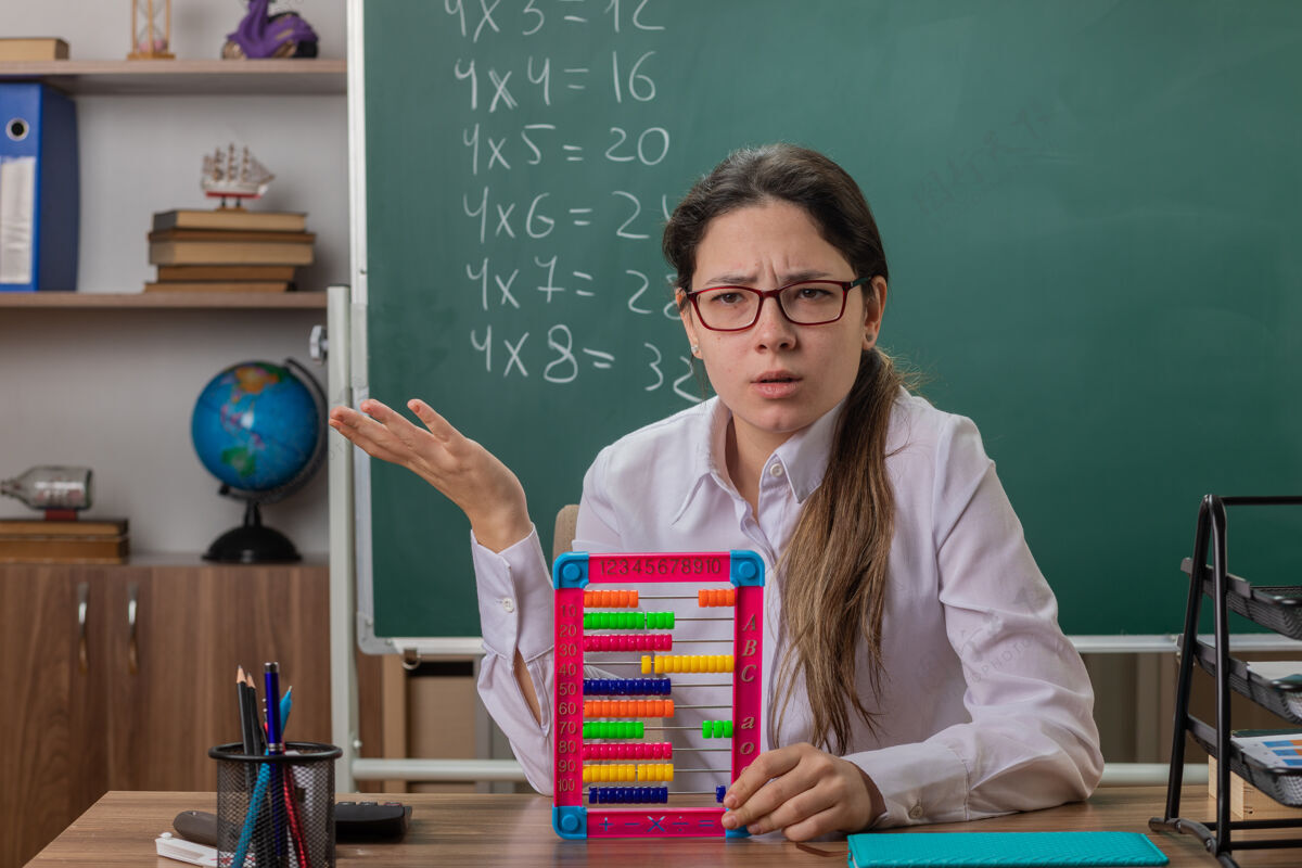 课戴着眼镜的年轻女教师坐在教室的黑板前 拿着账单 一脸困惑困惑书桌年轻
