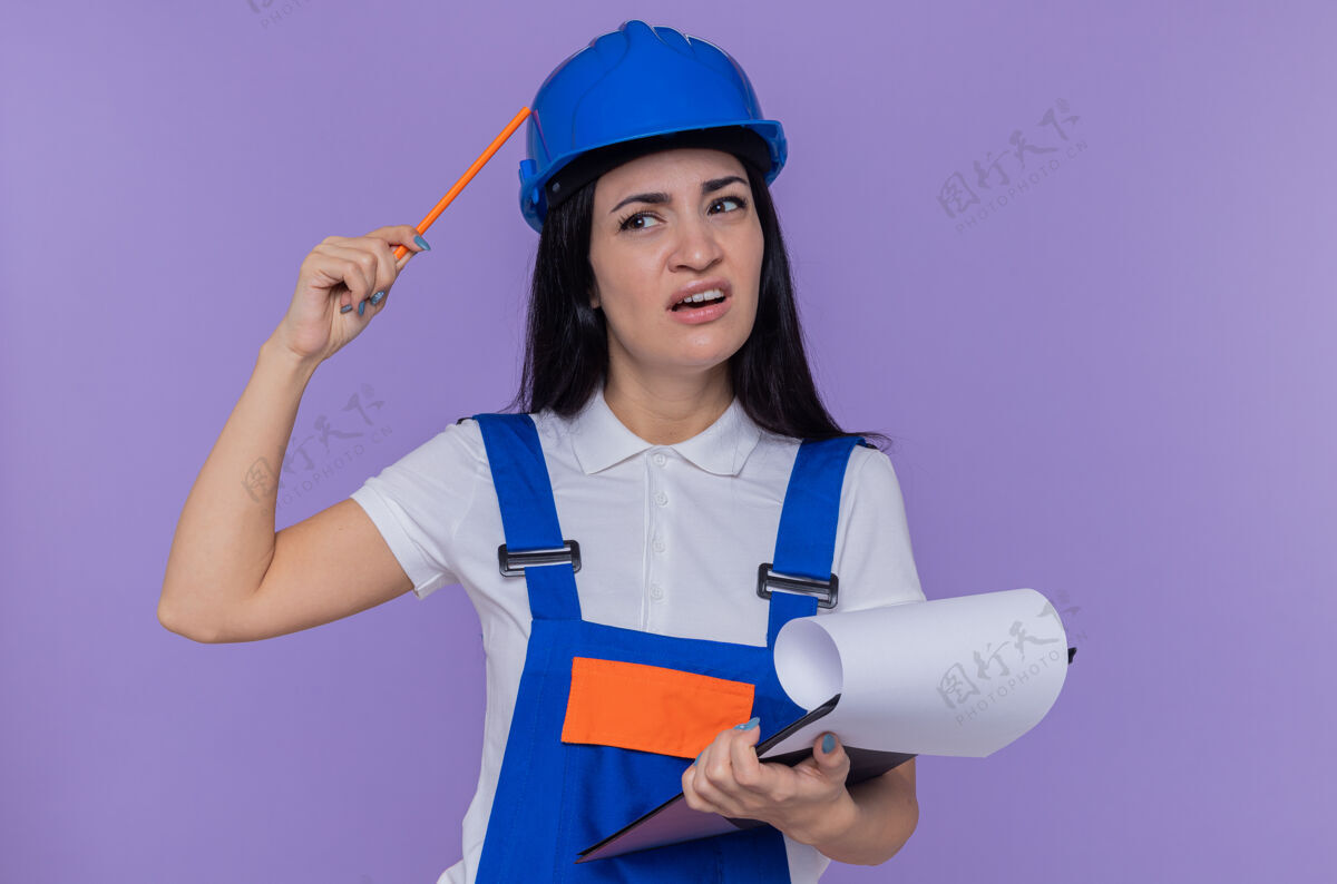 年轻一个穿着建筑制服 戴着安全帽 拿着写字板和铅笔的年轻建筑妇女站在紫色的墙上 困惑地一边看一边挠头建设者抓着放在一边