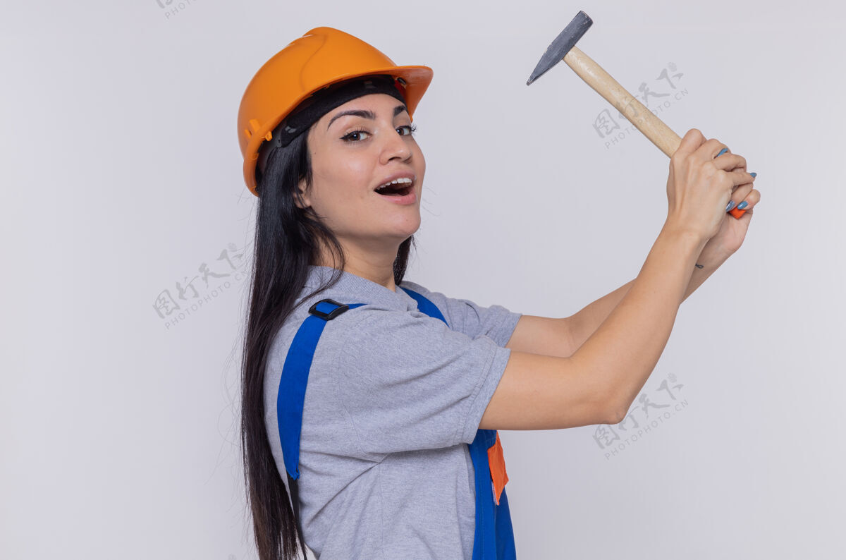 自信身穿施工制服 头戴安全帽 手持铁锤的年轻建筑工人自信地微笑着站在白色墙壁上看着前方建筑锤子微笑