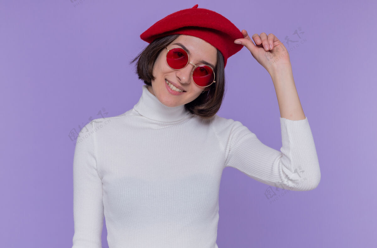触摸快乐积极的年轻女子 短发 白色高领毛衣 头戴贝雷帽 戴着红色太阳镜 面带微笑 站在蓝色的墙上 轻抚着贝雷帽积极高领毛衣头发