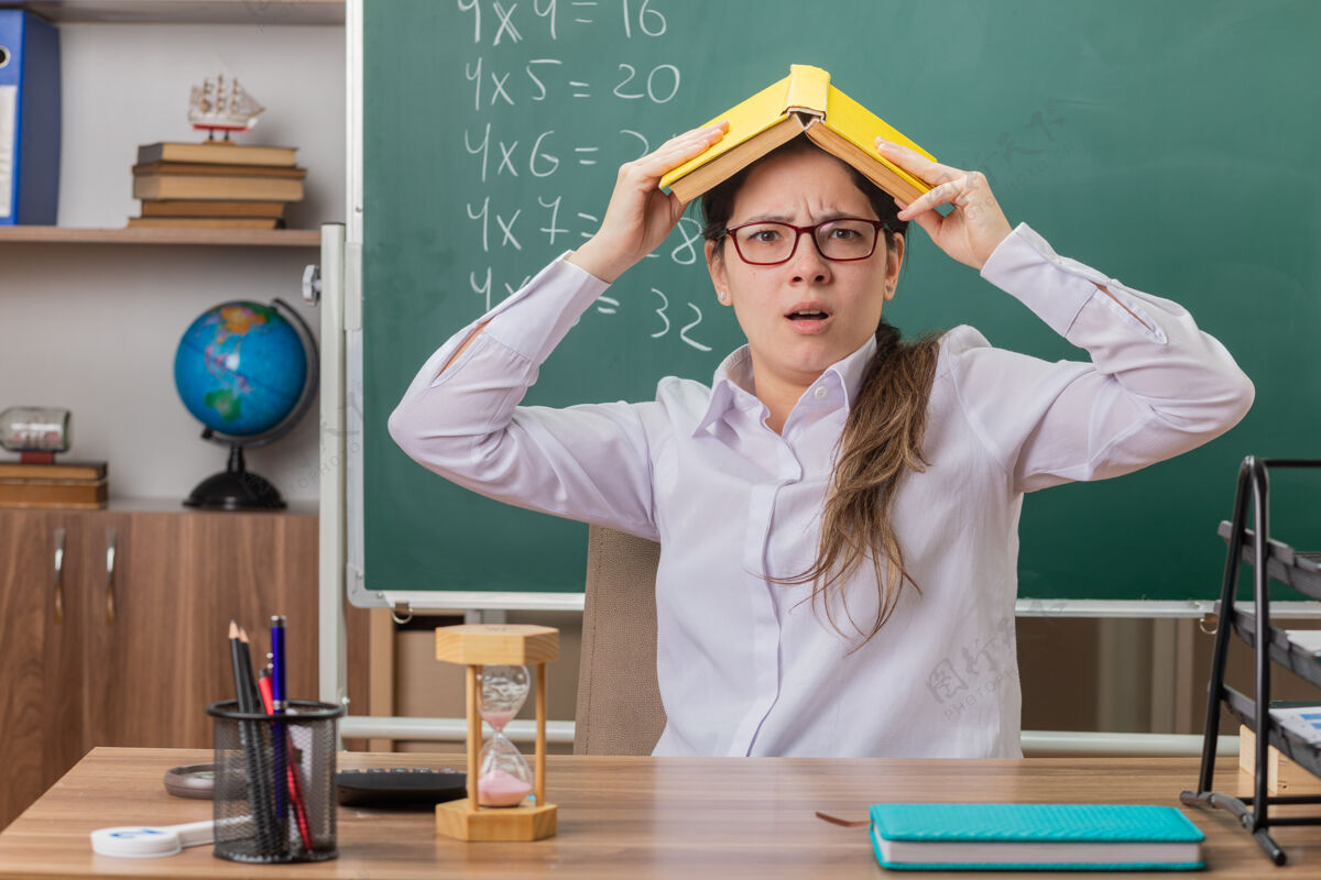 眼镜年轻的女老师戴着眼镜 头上抱着书 坐在教室黑板前的课桌旁 看上去又累又烦女人穿烦