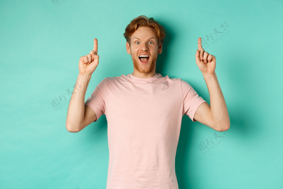 休闲穿着t恤的红发男人 手指朝上 敬畏地盯着镜头 展示着广告 站在绿松石色的背景上脸学生胡须