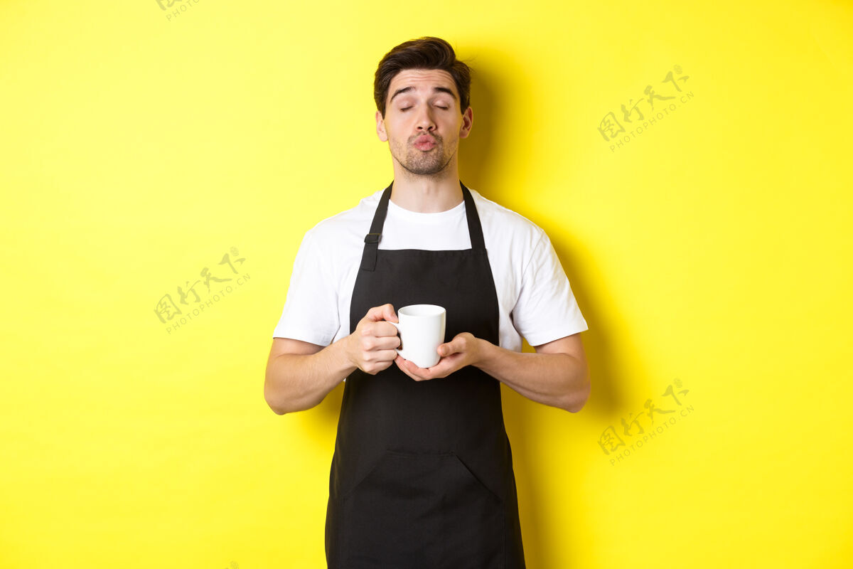 工作室一个穿着黑色围裙的男人端着一杯咖啡 站在黄色的背景上等待亲吻咖啡师模特围裙