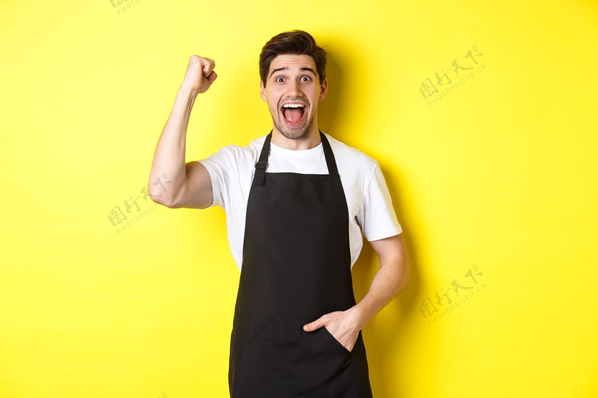 厨师兴高采烈的卖家在做拳头泵 欢欣鼓舞 得意洋洋 站在黄色背景下的黑色围裙上时尚自信男人