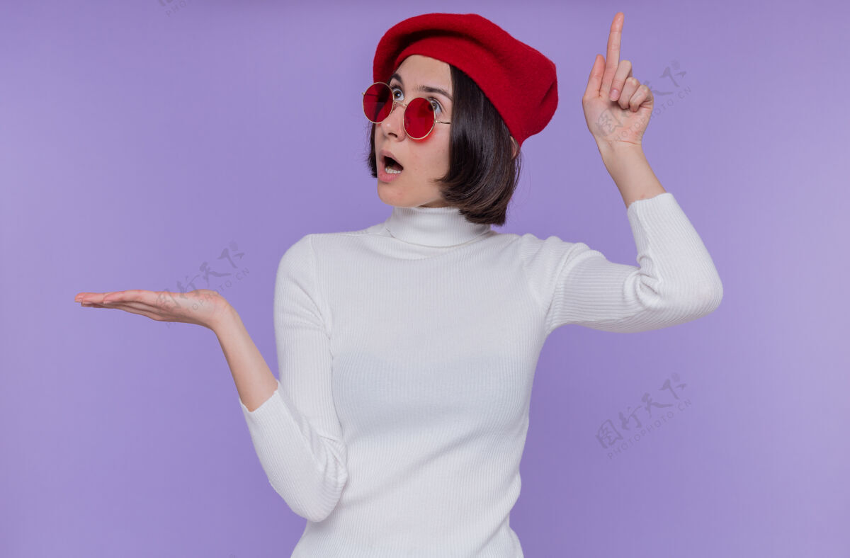 手臂一个短发 穿着白色高领毛衣 头戴贝雷帽 戴着红色太阳镜的年轻女子 看起来很惊讶 她伸出一只胳膊 食指朝上 站在蓝色的墙上介绍短裤惊喜