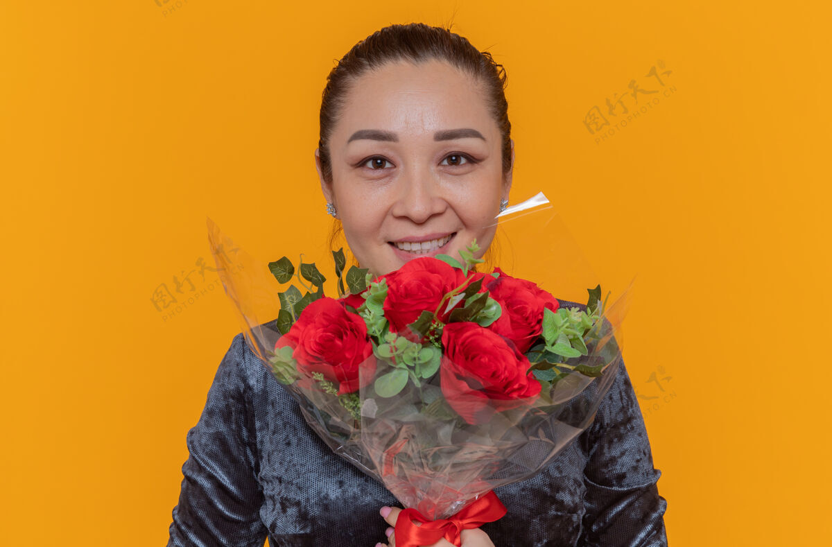 玫瑰快乐的亚洲女人捧着一束红玫瑰望着前方微笑着站在橙色的墙上欢庆国际妇女节庆祝举办微笑