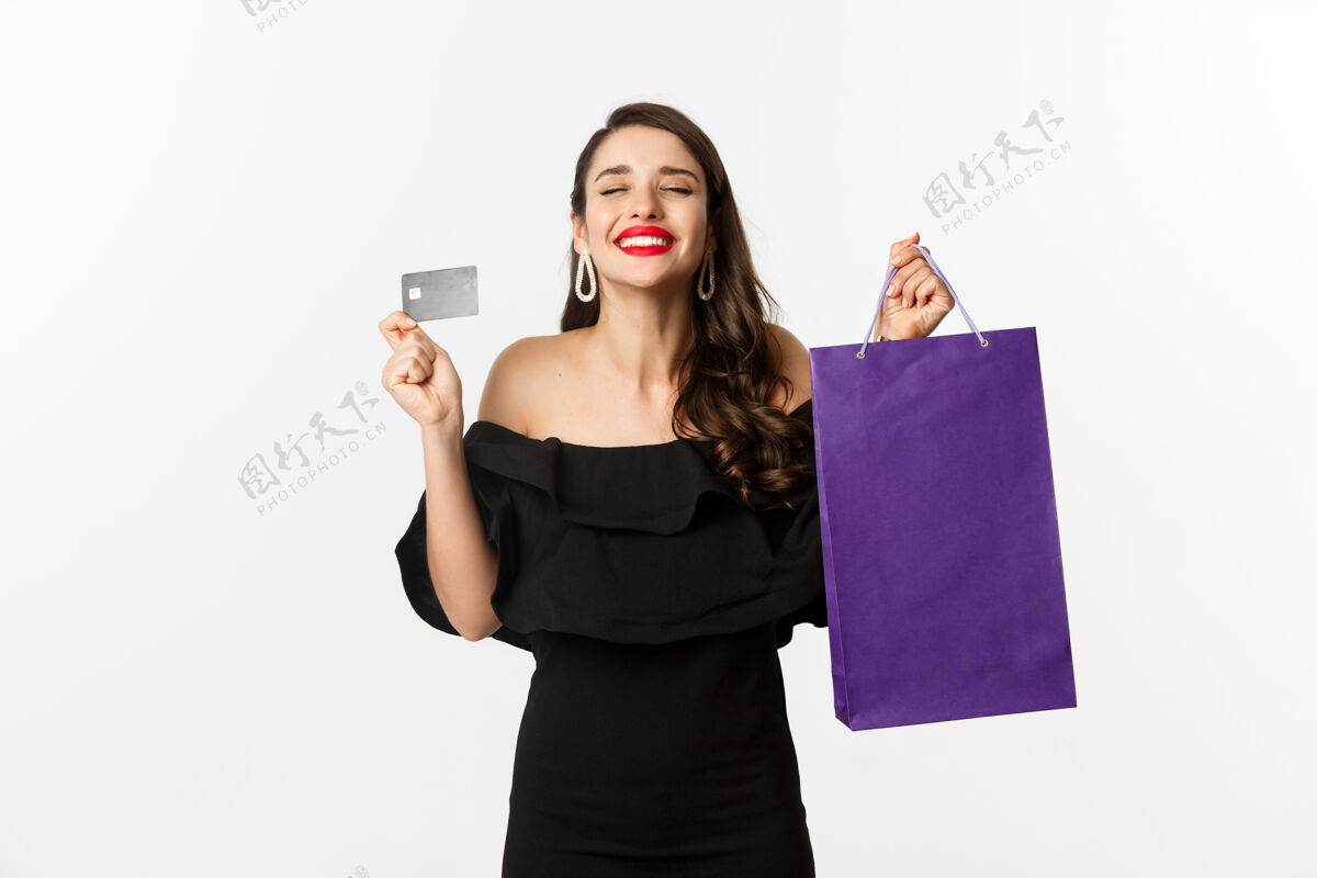 时尚时尚的年轻女子穿着黑色衣服去购物 拿着包和信用卡 高兴地微笑着 站在白色的背景下表情年轻服饰