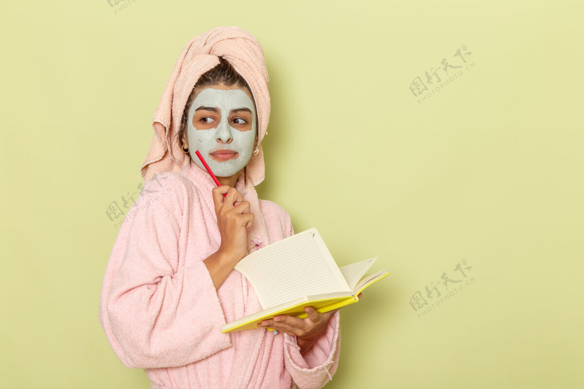 年轻女性正面图穿着粉红色浴袍的年轻女性在浅绿色的表面写下笔记羽绒服洗澡浴袍