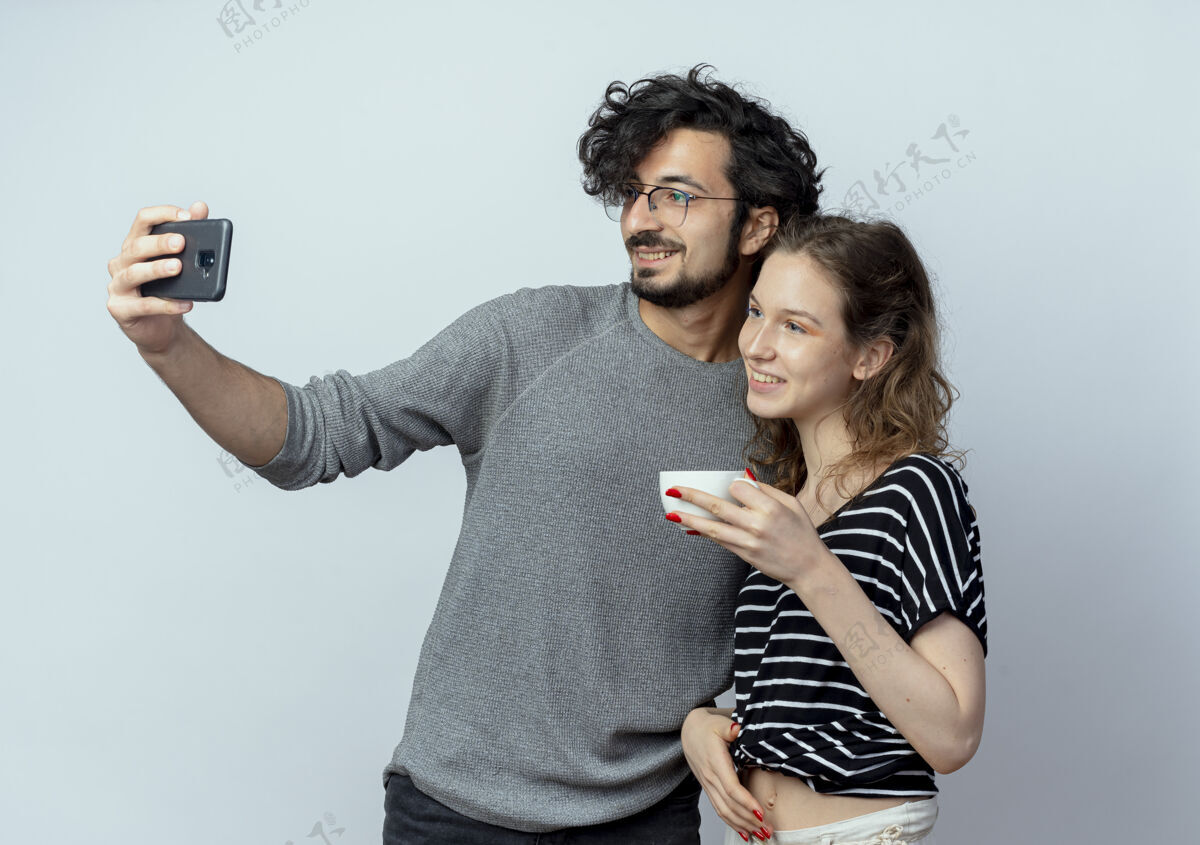 女人一对年轻的男女 快乐的男人用智能手机拍下了他们的照片 而他的女朋友站在他旁边 隔着白墙喝着咖啡拿着站着手机