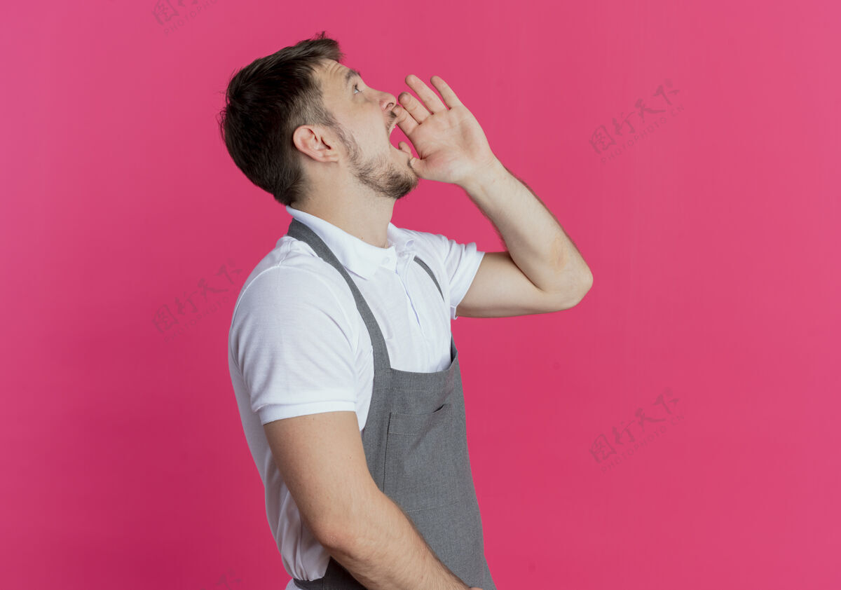 靠近围裙上的理发师站在粉红色的墙上 用手靠近嘴巴喊着或叫着别人有人围裙理发师