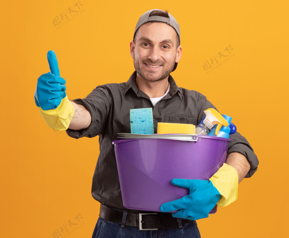 穿着年轻人穿着休闲服 戴着橡胶手套 手里拿着带清洁工具的水桶 面带微笑 自信地竖起大拇指站在橙色的墙上自信拇指清洁