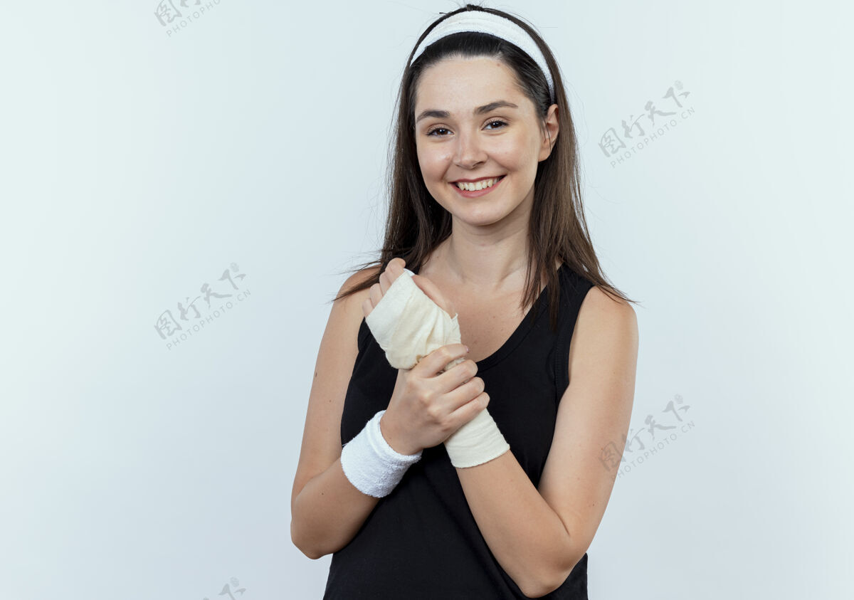健康戴着头巾的年轻健身女士站在白墙上 面带微笑地抚摸着缠着绷带的手腕手腕触摸头带
