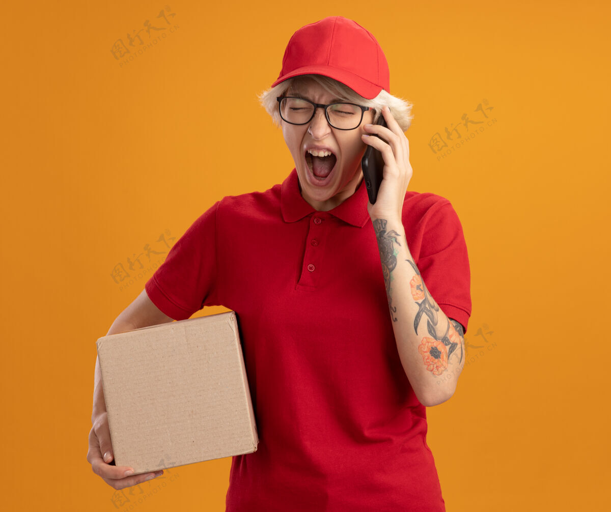 帽子身穿红色制服 戴着帽子 戴着眼镜 拿着纸板箱的年轻送货员站在橘红色的墙上 一边用手机说话 一边用咄咄逼人的表情大喊大叫表情同时送货