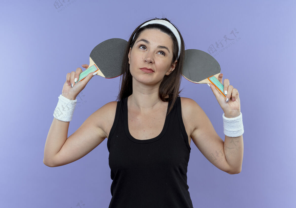 女子戴着头巾拿着球拍准备打网球的年轻健身女士站在蓝色的墙上困惑地仰望着举行拼图年轻