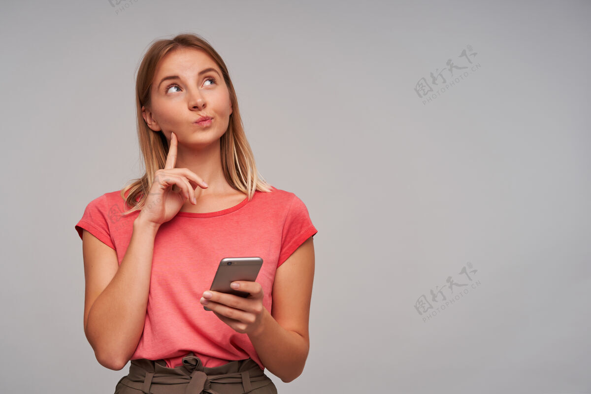 奇迹可爱的 好奇的金发女孩的肖像穿着粉红色的t恤和棕色的裙子拿着手机 摸着她的脸颊在灰色的墙上的复印空间看着右上角装装模型脸