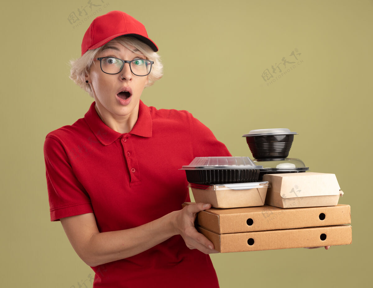 拿着年轻的送货员身穿红色制服 戴着眼镜 手里拿着披萨盒和食品包 站在绿色的墙上 既惊讶又担心盒子食物帽子