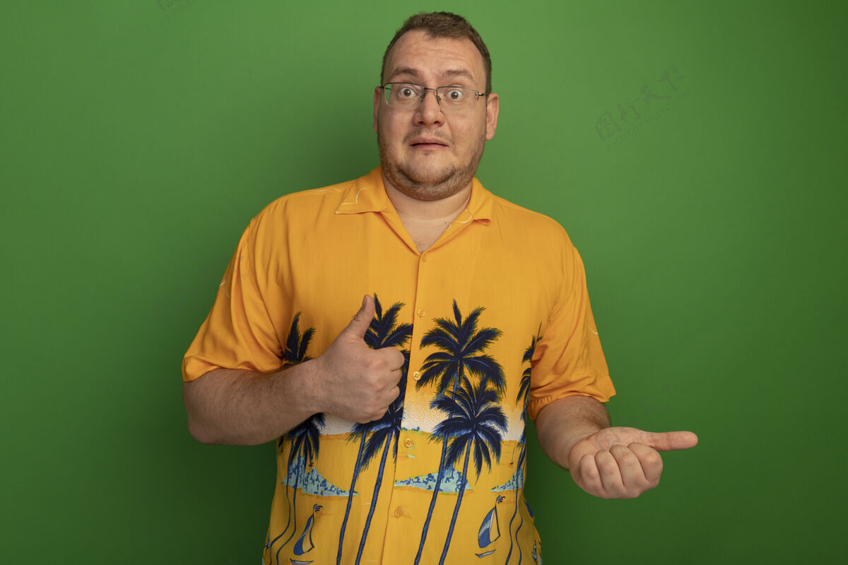 站起来一个戴着眼镜和夏威夷衬衫的男人站在绿色的墙上竖起大拇指男人困惑拇指