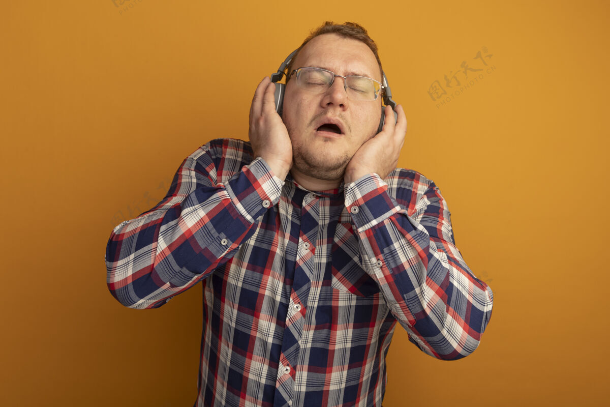 男人戴着眼镜 穿着格子衬衫 戴着耳机的男人站在橙色的墙上欣赏音乐音乐眼镜耳机