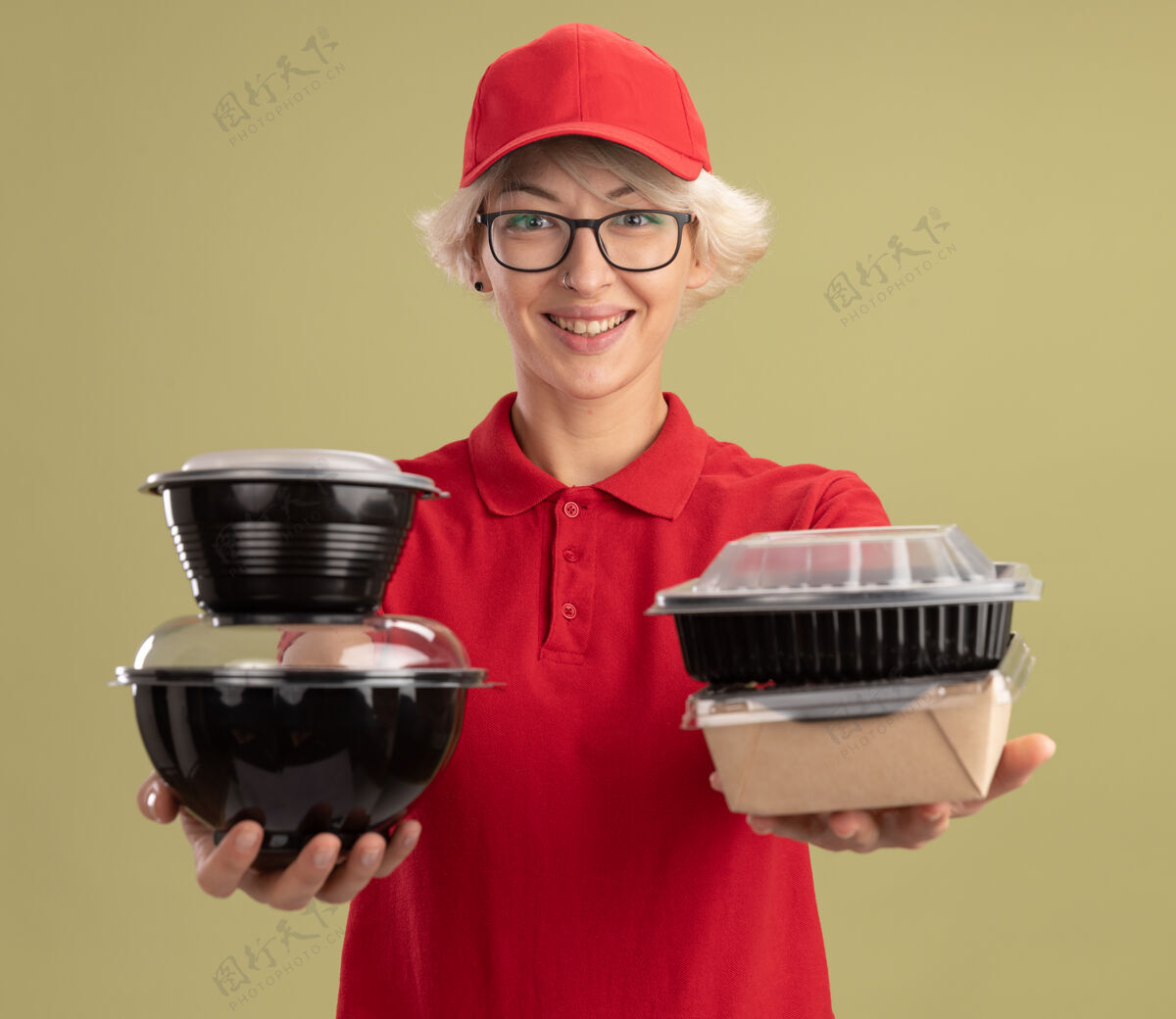 帽子年轻的送货员身穿红色制服 戴着眼镜 面带微笑地拿着食品包站在绿色的墙上盒子戴着站着
