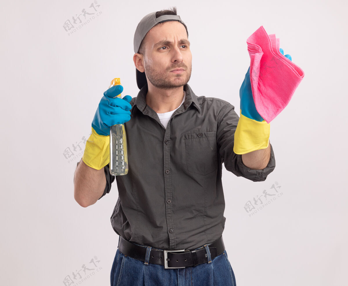 男人年轻的清洁工穿着便服 戴着橡胶手套 戴着帽子 手里拿着清洁剂和抹布 站在白色的墙上 严肃地看着旁边手套帽子清洁
