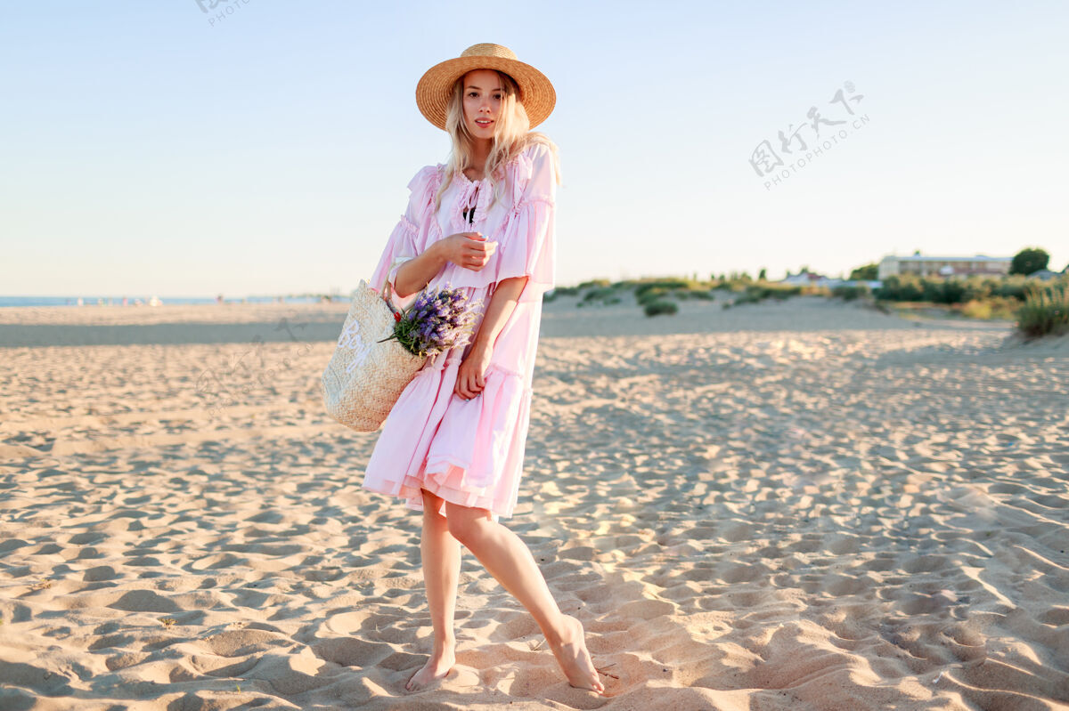 海滩金发女孩穿着可爱的粉色裙子在沙滩上跳舞 手里拿着草包和鲜花美丽优雅风