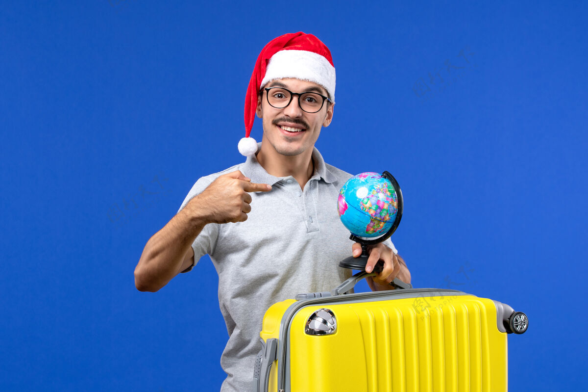球员正面图：年轻男性提着黄色包和地球仪在蓝色书桌上度假旅行人飞机积极