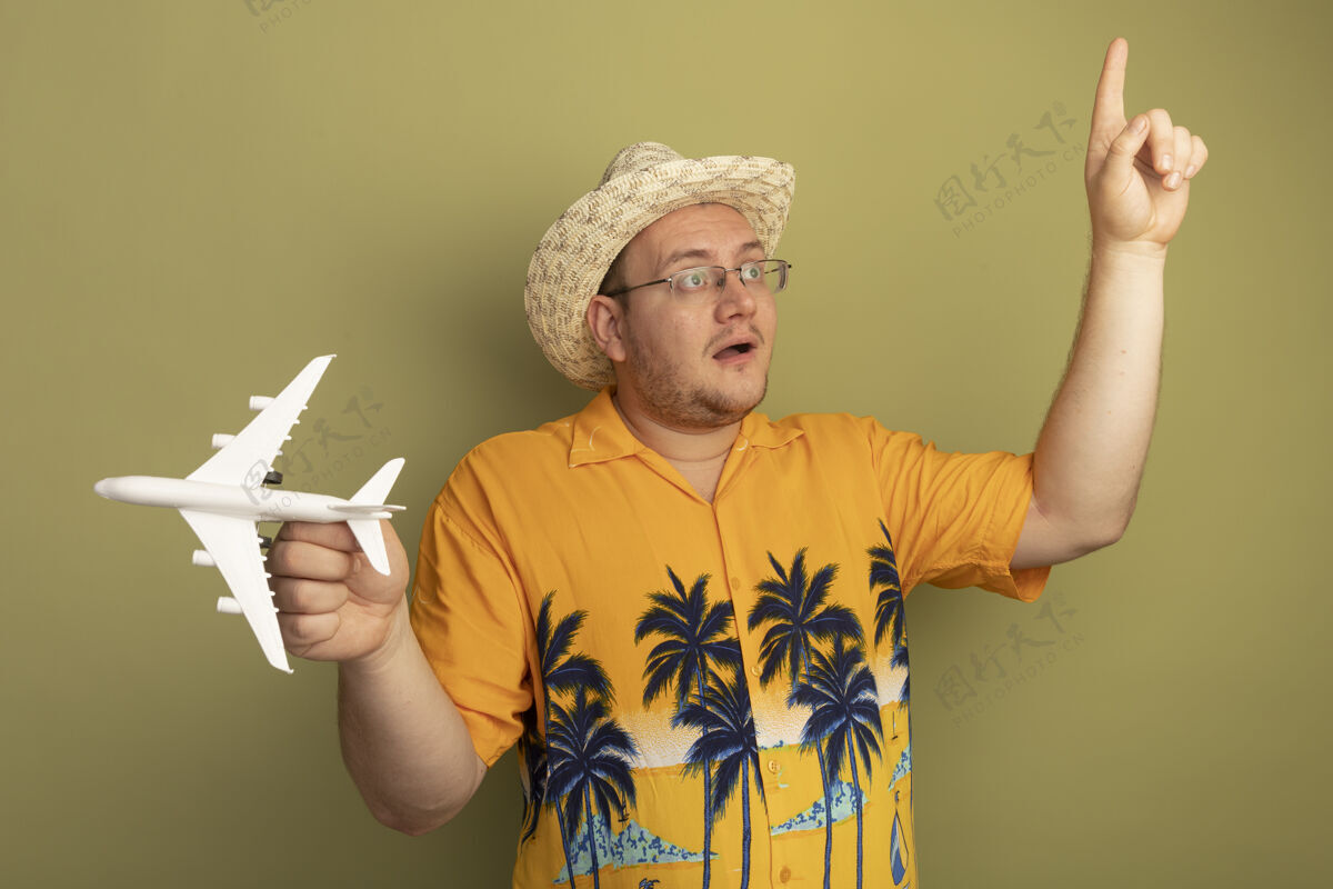 立场戴着橘色衬衫戴着夏天帽子戴着玩具飞机的戴着眼镜的男人站在绿色的墙上 一边看一边露出食指 惊讶而快乐夏天眼镜目录