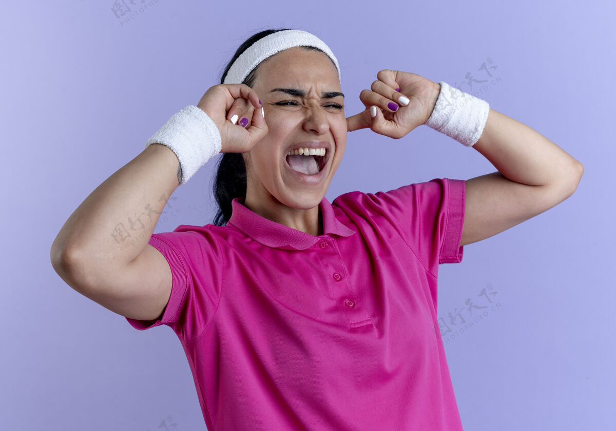 运动年轻恼怒的高加索运动女性戴着头带和腕带闭上耳朵看着紫色空间与复制空间隔离的一面头带恼火看