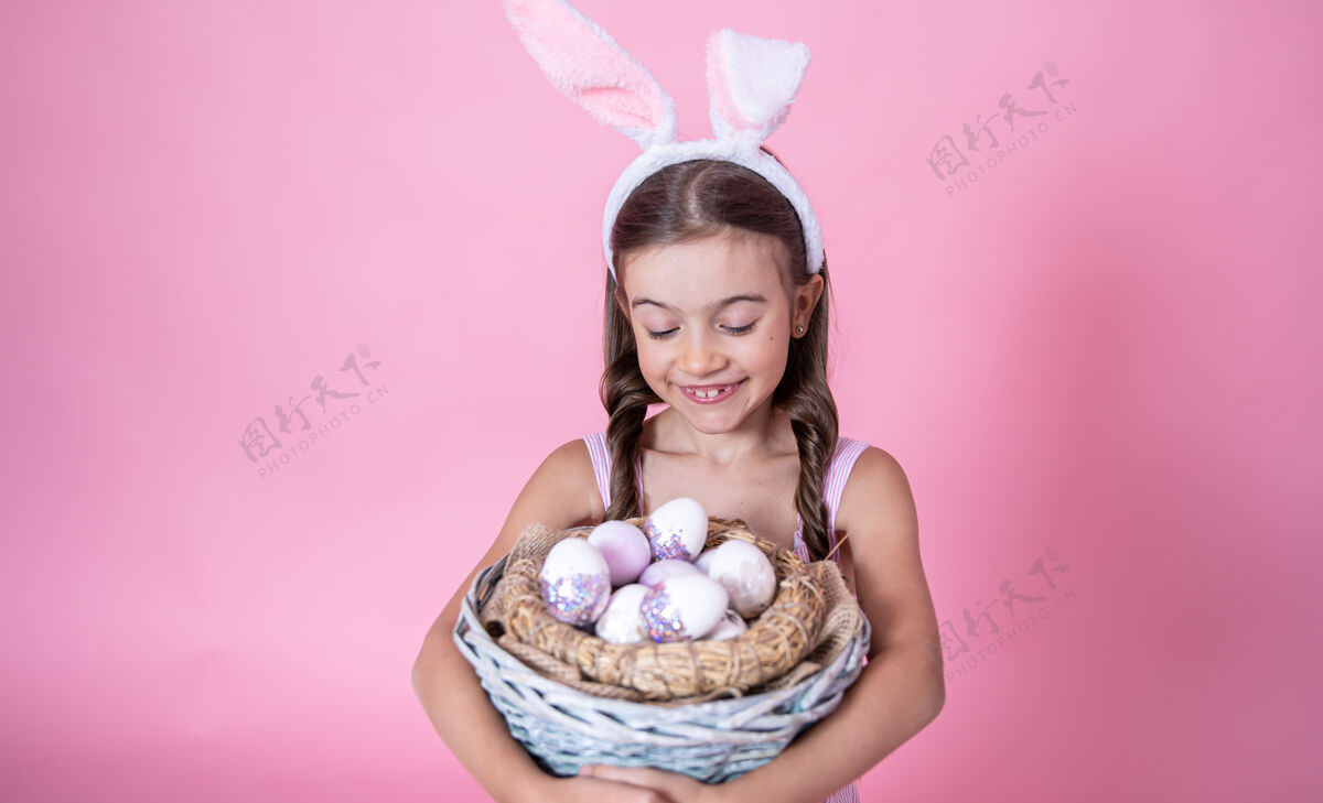 姿势带着复活节兔子耳朵的小女孩摆姿势 手里拿着一个篮子 篮子里放着粉红色的复活节彩蛋模型装饰篮子