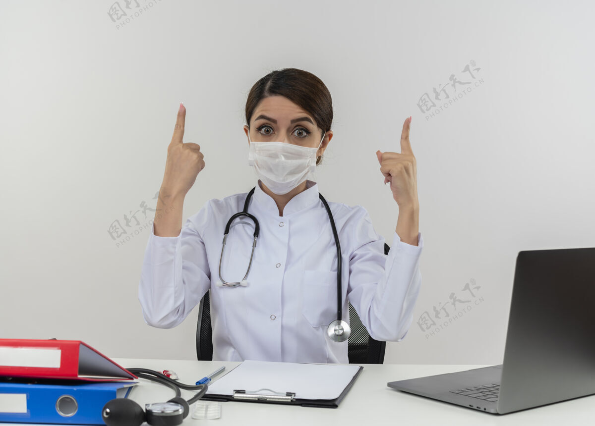 印象年轻的女医生穿着医用长袍 戴着听诊器和医用面罩 坐在办公桌旁 手持医疗工具和笔记本电脑 孤零零地站在白色的墙上医疗长袍指着