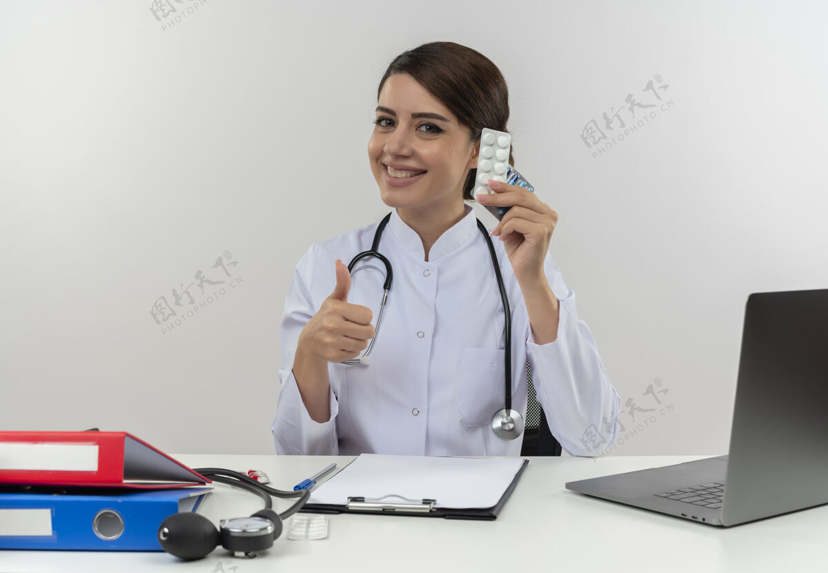 穿面带微笑的年轻女医生穿着医用长袍 听诊器坐在办公桌旁 手里拿着医疗工具和手提电脑 手里拿着药品 在白色的墙上孤立地竖起大拇指微笑长袍拇指
