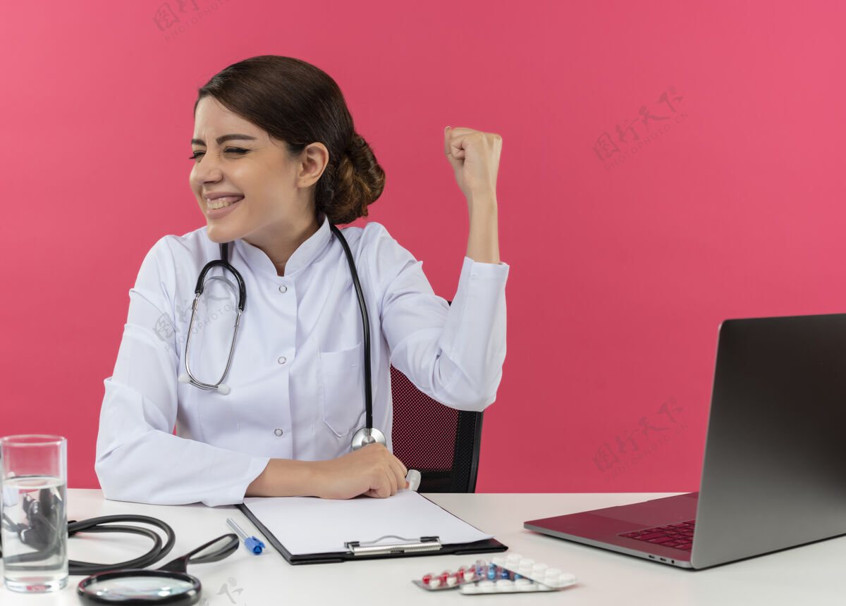 年轻快乐的年轻女医生穿着医用长袍和听诊器坐在办公桌旁 手里拿着医疗工具和笔记本电脑 头转向一边做着“是”的手势 眼睛紧闭 隔离在粉红色的墙上笔记本电脑坐着医疗