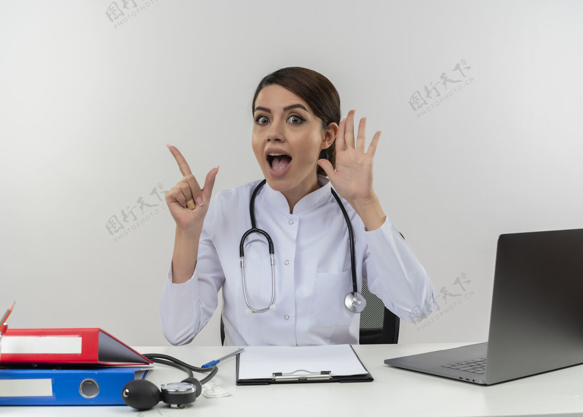 靠近年轻的女医生穿着医用长袍和听诊器坐在办公桌旁 手里拿着医疗工具和笔记本电脑 手靠近耳朵 手指被隔离在白色的墙上笔记本电脑手医疗
