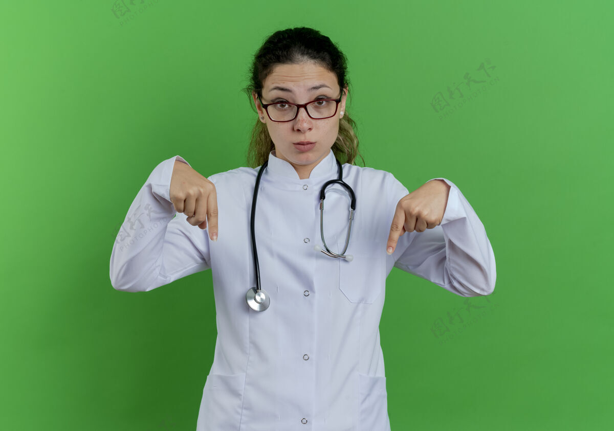 指向年轻的女医生穿着医用长袍 戴着听诊器 戴着眼镜 孤独地指着绿色的墙上看医生长袍