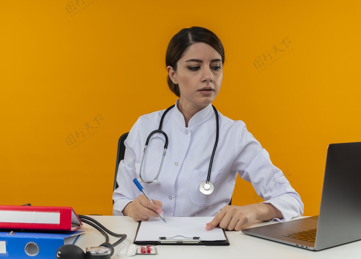 医生专注的年轻女医生穿着医用长袍和听诊器坐在黄色墙上的桌子上专心剪贴板听诊器