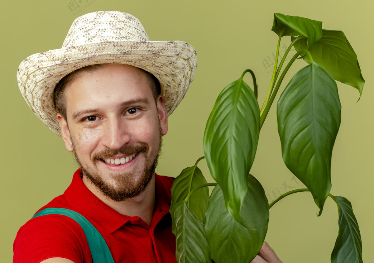 植物特写镜头中的快乐年轻英俊的斯拉夫园丁制服和帽子举行植物隔离橄榄绿墙看特写斯拉夫