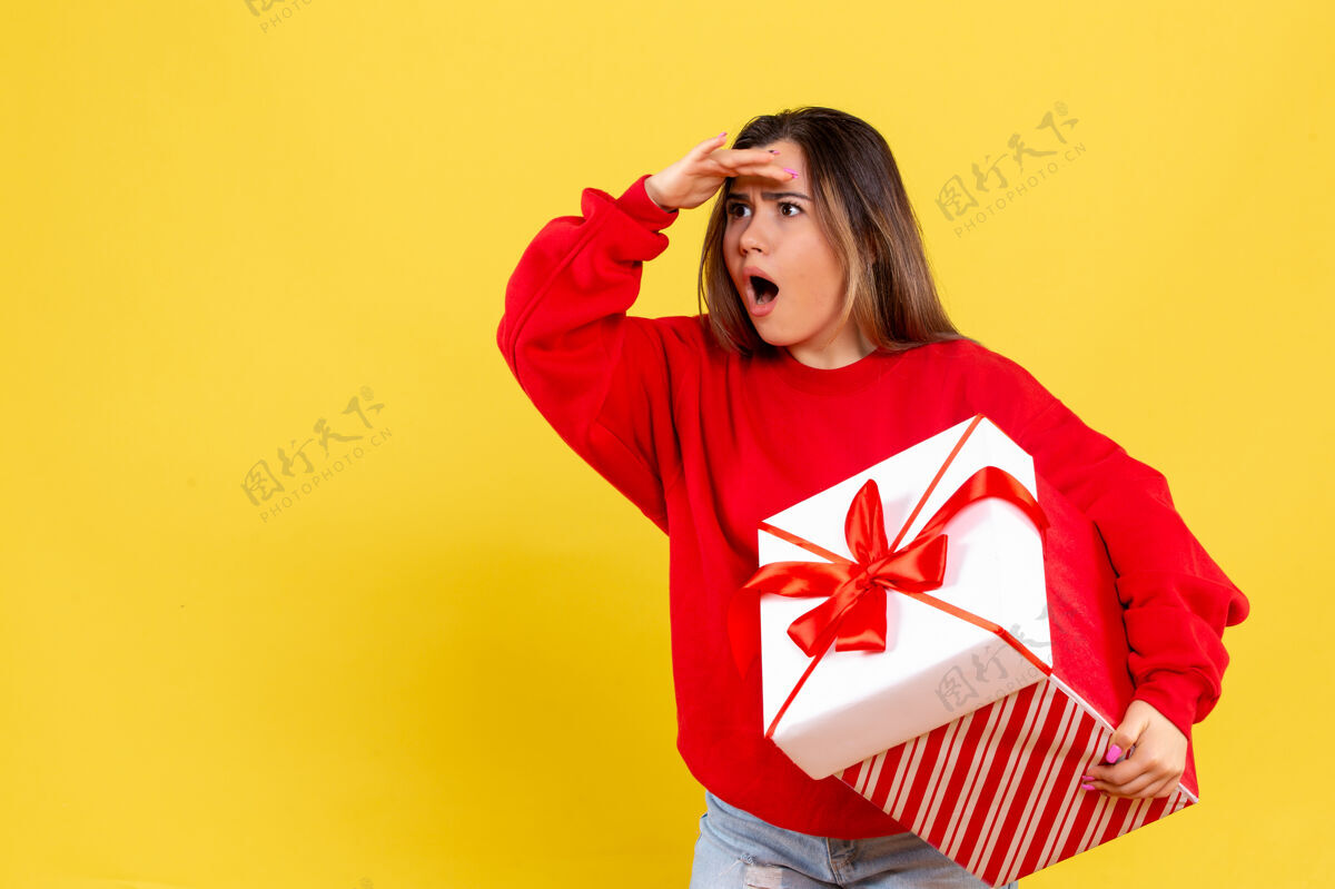 肖像前视图年轻女子手持圣诞礼物惊讶地看着黄色背景上的远处礼物距离成人