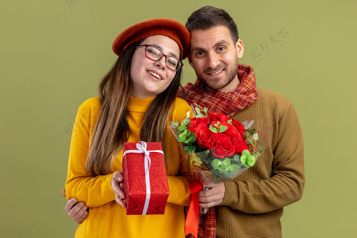 年轻年轻漂亮的夫妇幸福的戴贝雷帽的女人拿着礼物 男人拿着一束红玫瑰看着镜头微笑着 一起欢庆情人节站在绿色的墙上情侣贝雷帽情人节