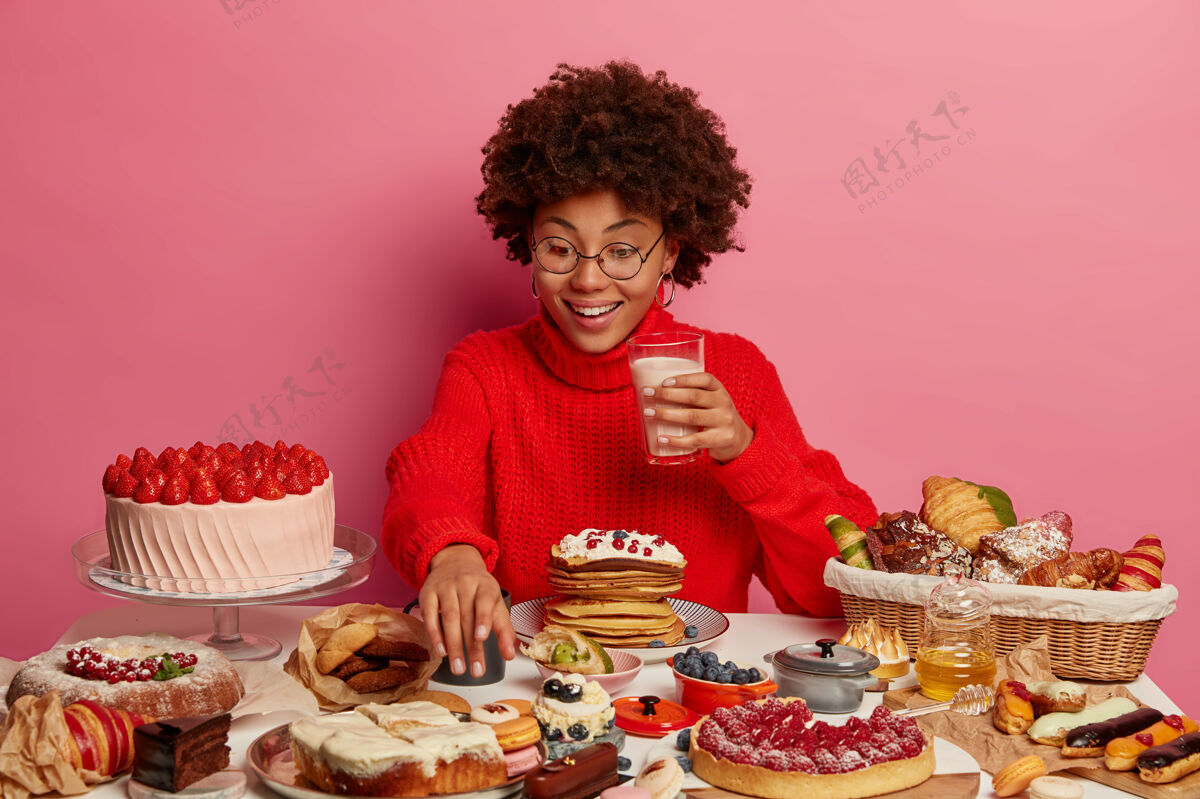 人开朗的非洲女人伸手去拿美味的甜点 拿着一杯牛奶 吃着蛋糕 周围都是垃圾食品 戴着眼镜和红色毛衣 不能拒绝甜食美味奶油味道