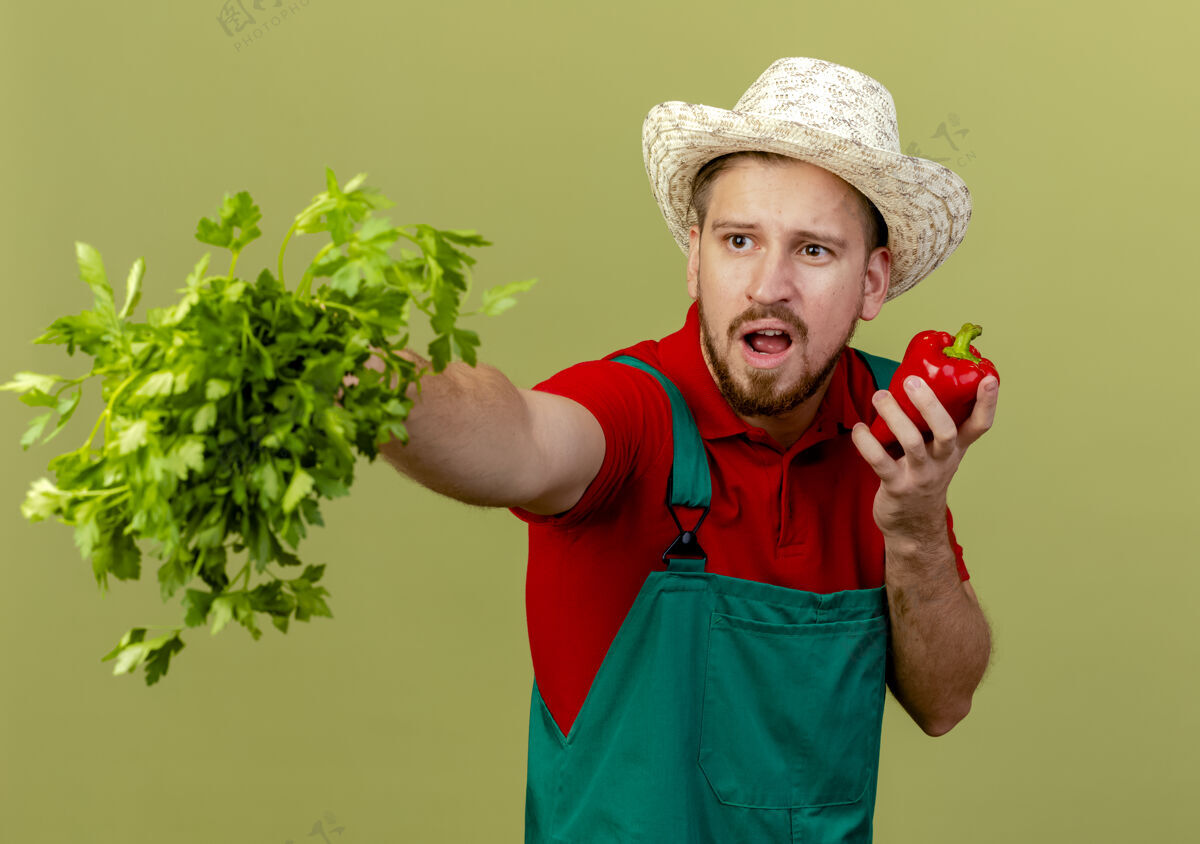 帅气年轻帅气的斯拉夫园丁穿着制服 戴着帽子 伸出香菜 看着一边拿着辣椒隔离在橄榄绿的墙上壁板制服帽子