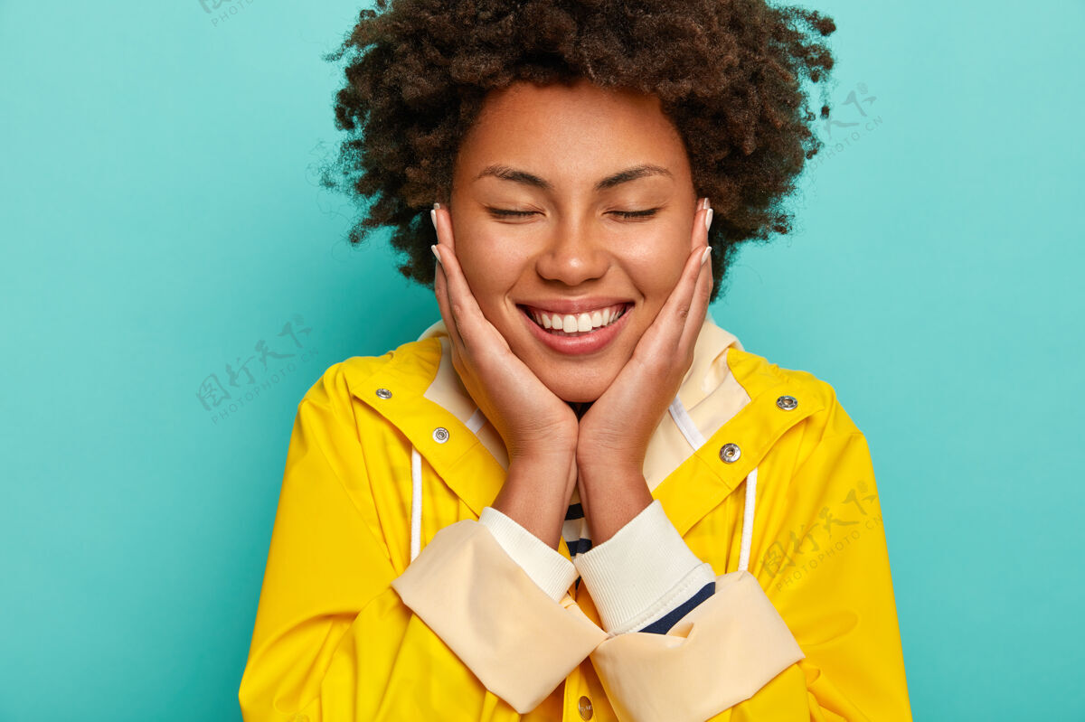 女人快乐开朗的非洲裔美国妇女的头像抚摸脸颊 闭上眼睛 享受愉快的时刻 穿着黄色雨衣 隔离在蓝色的背景上快乐人快乐