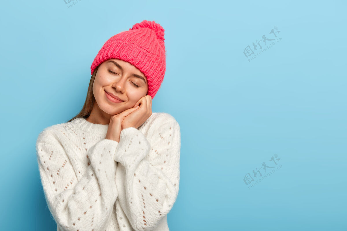 帽子温柔的年轻女子闭上眼睛 歪着头 想象着一些令人愉快的事情 戴着粉色的帽子 穿着冬衣 在蓝色的背景下摆姿势年轻想象肖像