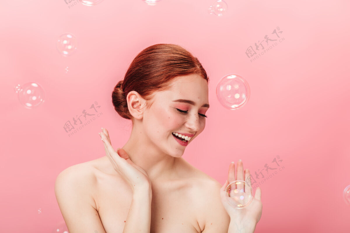 姜黄色无忧无虑的漂亮女孩摆出肥皂泡的姿势摄影棚拍摄的迷人的姜黄色女人在粉色背景下微笑红发表情欢呼