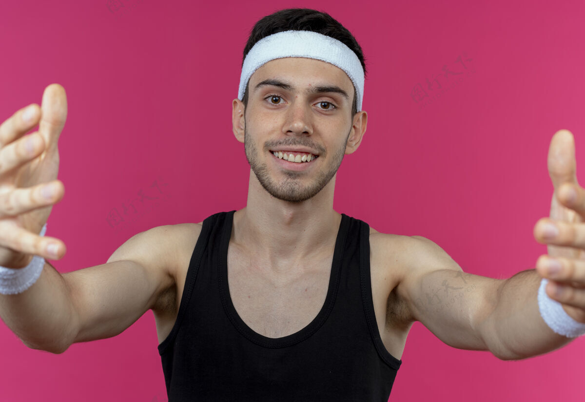 手戴着头巾的年轻运动型男子做着欢迎的手势 双手微笑着越过粉色站立手势年轻