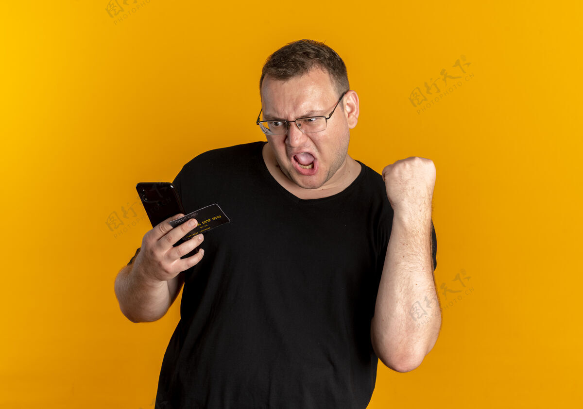 穿戴眼镜的超重男子身穿黑色t恤手持智能手机握紧拳头大喊橙色超重拳头男人
