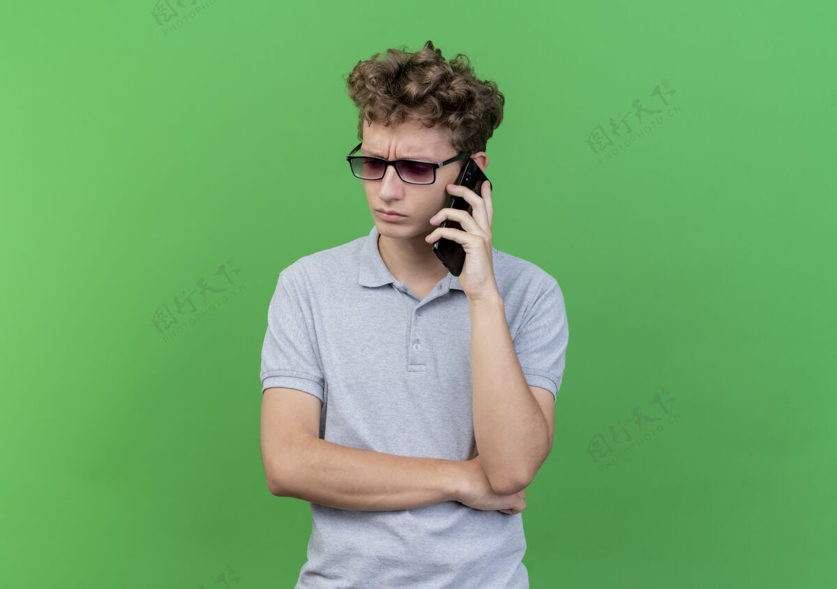 而戴着黑眼镜 身穿灰色马球衫的年轻人一边迷茫地看着一旁 一边用绿色手机交谈年轻穿衬衫