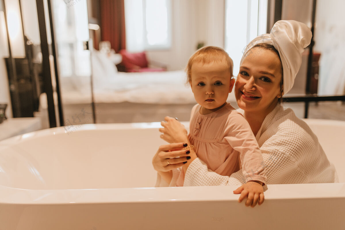 女人妈妈和小女儿坐在白色浴缸里微笑着看着镜头浴室毛巾浴袍