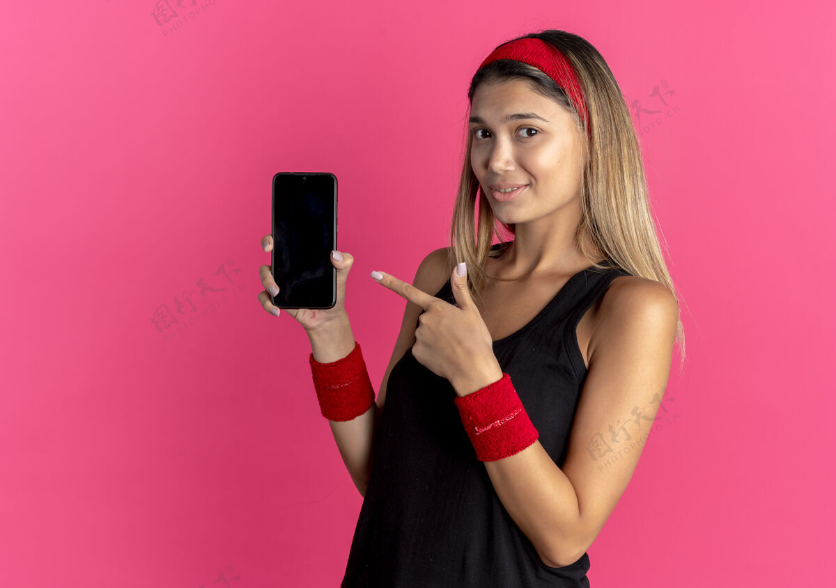 年轻身穿黑色运动服 头戴红色头带的年轻健身女孩展示着智能手机指针 手指对着它微笑着越过粉色黑色红色表演