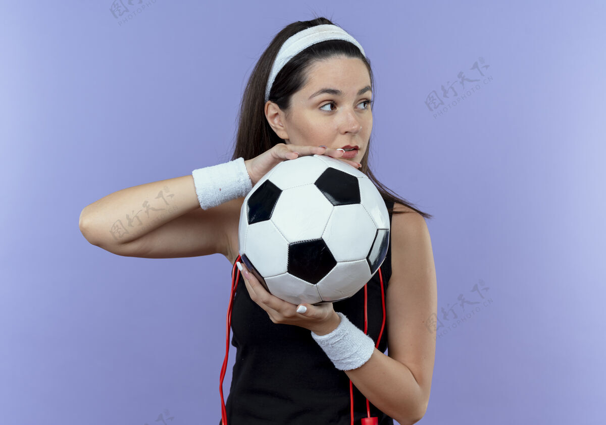 站戴着头巾 脖子上系着跳绳 手持足球 神情自信地站在蓝色墙壁上的年轻健身女士足球球看