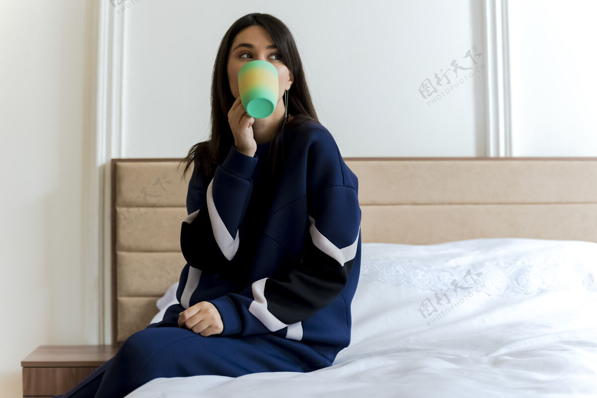 咖啡年轻漂亮的白种女人坐在卧室的床上 手放在腿上 一边看一边喝咖啡黑发床腿